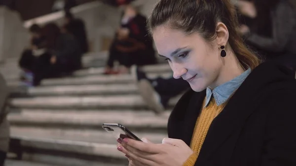 Jovem morena sentada na escada na multidão e usando smartphone. Menina olhando ao redor e sorrindo . — Fotografia de Stock