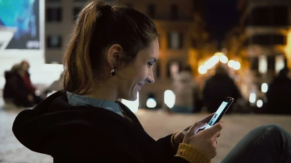 Mujer morena atractiva sentada en el centro de la ciudad por la noche y mirando la foto en el teléfono inteligente con tecnología de pantalla táctil . —  Fotos de Stock