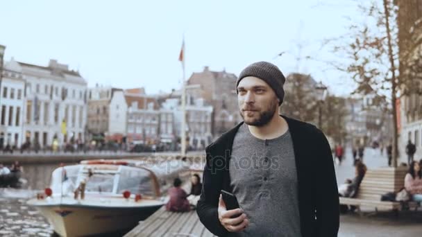 4k tourist man walking with smartphone, amsterdam. lässiger gutaussehender bärtiger lächelnder Mann spaziert in der Nähe eines Flussufers. — Stockvideo