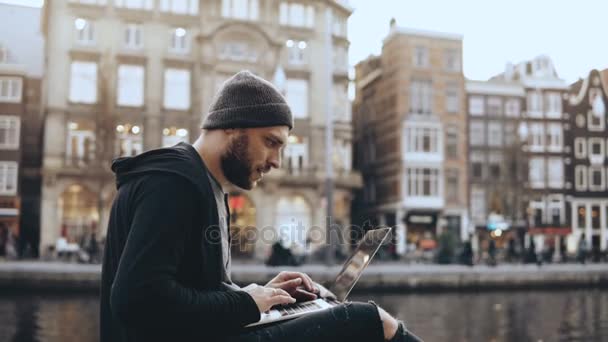 4k affärsman som sitter med en bärbar dator nära floden. Kreativ konst arbetaren glad och framgångsrik. Amsterdam, Nederländerna. — Stockvideo