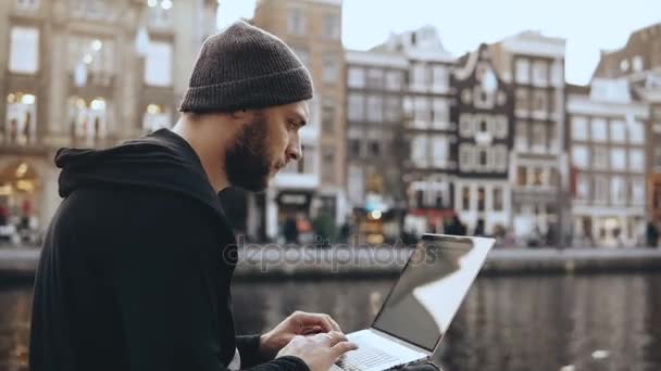 4К Взрослый бизнесмен 30-х годов пользуется ноутбуком на открытом воздухе. Сконцентрированный человек, работающий онлайн. Мобильное рабочее место. Амстердам старый город . — стоковое видео