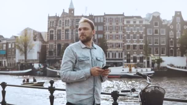 4K Hombre casual mirando a su alrededor con teléfono inteligente. Casual guapo hombre barbudo confundido con bicicleta cerca de un terraplén del río . — Vídeo de stock