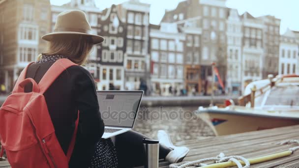 4K 30s empresaria feliz que trabaja con el ordenador portátil. Amsterdam. Señora con mochila roja se sienta en el hermoso terraplén del río . — Vídeos de Stock