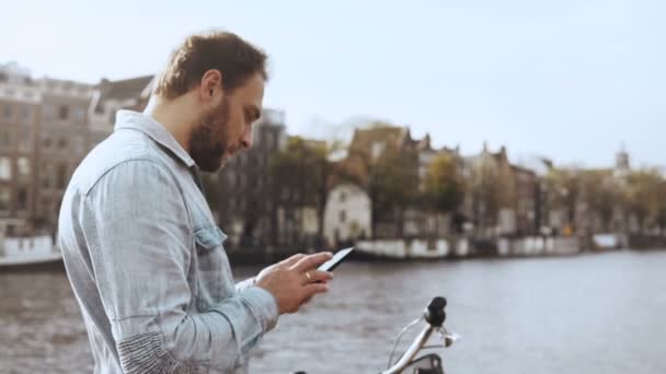 Homem de negócios europeu 4K a enviar mensagens numa ponte fluvial. Homem barbudo adulto com bicicleta em camisa azul trabalhando em escritório móvel . — Vídeo de Stock