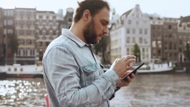 4K homme d'affaires caucasien textos près de la rivière. Plan panoramique. 30s sérieux barbu homme en chemise bleue envoyer des messages électroniques . — Video