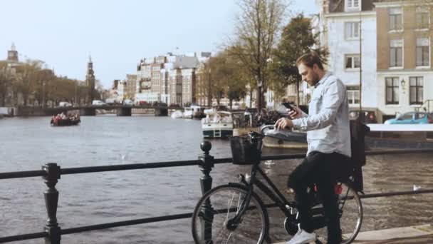 4k 欧洲人自行车在河桥梁。在智能手机上休闲时尚的男性类型, 环顾四周享受美景. — 图库视频影像