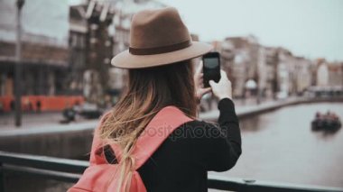 Şapkalı genç kız doğal nehir fotoğraflarını çeker. Kız uzun saçlı ve kırmızı sırt çantası onun blog resimleri paylaşma. 4k.