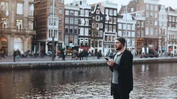 4K Casual Caucasian man in hat using smartphone. Handsome bearded creative businessman texting on a river embankment. — Stock Video