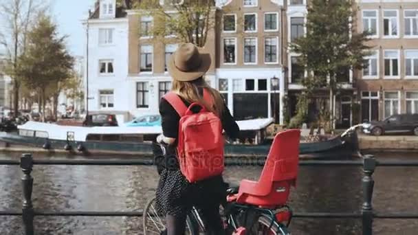 Linda chica se baja de una bicicleta, mira a su alrededor. Joven blogger de estilo de vida con bicicleta y flores en el pintoresco puente. 4K — Vídeo de stock