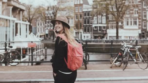 Uma mulher muito caucasiana a atravessar uma rua de bridge. 4K. Jovem viajante feminino na moda com mochila falando no telefone . — Vídeo de Stock