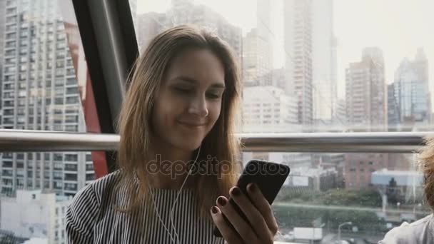 4k πορτρέτο της Ευρωπαϊκή κορίτσι στη Νέα Υόρκη τραμ αέρα. Όμορφη κυρία με smartphone και ακουστικά στο Μανχάταν τελεφερίκ. — Αρχείο Βίντεο