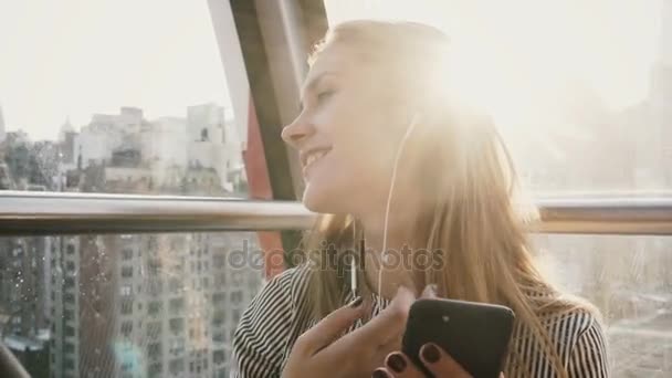 4K Menina caucasiana feliz em Island Tramway, NY. Menina bonita com smartphone e fones de ouvido em um funicular. Sinal de luz solar . — Vídeo de Stock