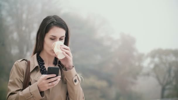 Junge schöne Frau, die im nebligen Morgen im Park steht und das Smartphone benutzt, Kaffee trinkt. — Stockvideo