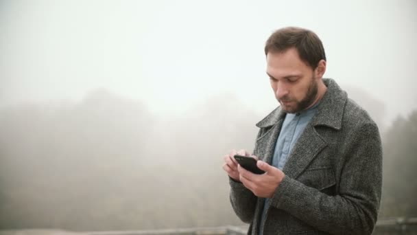 Retrato de un joven guapo parado en el parque de niebla temprano en la mañana. Varón barbudo usando smartphone con pantalla táctil . — Vídeos de Stock