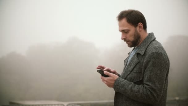 Joven hombre guapo de pie temprano en la mañana brumosa en el parque. Varón barbudo usando el smartphone con pantalla táctil . — Vídeos de Stock