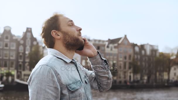 Heureux homme européen avec vélo parlant au téléphone. 4K Homme barbu adulte occasionnel parlant à un ami sur le quai de la rivière vieille ville . — Video