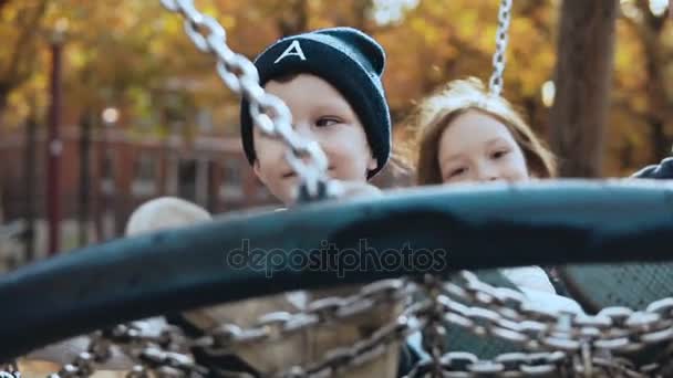 Dos niños pequeños balanceándose en un columpio de nido en el parque. Pequeños niños europeos felices mirando a la cámara sonriendo alegremente . — Vídeos de Stock