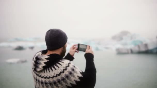 Ung snygg man står på stranden i Jokulsalon ice lagoon i Island och ta bilder på smartphone. — Stockvideo