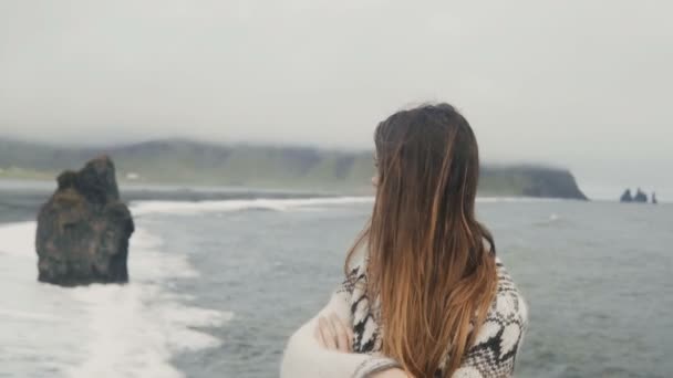 Joven hermosa mujer de pie en la playa negra, cerca de los dedos del pie Troll en Islandia y mirando a su alrededor en un día ventoso . — Vídeos de Stock