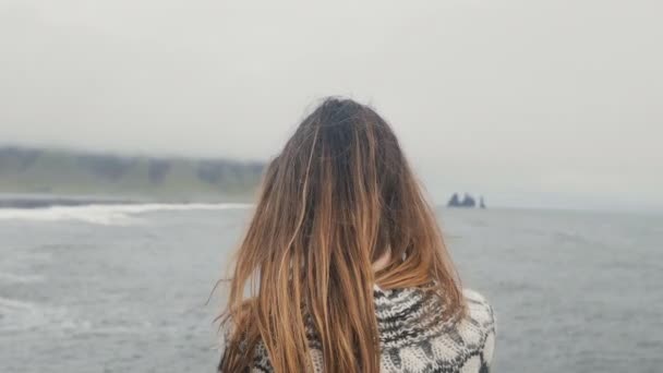 Вид сзади молодой брюнетки, стоящей на черном пляже и наслаждающейся прекрасным видом на скалы троллей в Исландии — стоковое видео