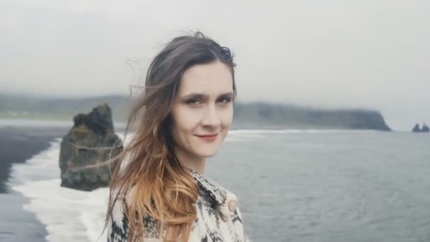 Portrait de jeune femme souriante debout sur la plage noire, orteils de troll en arrière-plan en Islande, regardant la caméra — Video