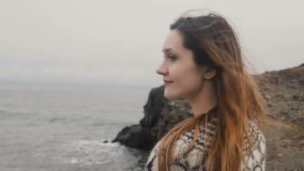 Porträt einer jungen glücklichen Frau, die am Ufer des Meeres steht und in die Kamera blickt, lächelt, die Haare wehen im Wind. — Stockvideo