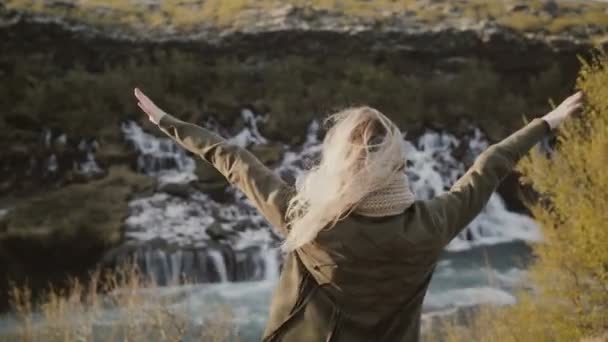 Свобода: молодая путешествующая женщина смотрит на водопады в Исландии и поднимает руки вверх, чувствуя себя счастливой . — стоковое видео