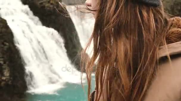Vista da vicino di una giovane donna in piedi vicino a una potente cascata in Islanda, i capelli che ondeggiano al vento . — Video Stock