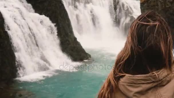 Зменшеному вигляді привабливих брюнетки самотньо стоїть і, дивлячись на потужний водоспад в Ісландії, волосся хвилі на вітер. — стокове відео