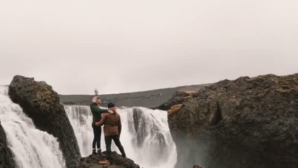 Vista posterior de la feliz pareja después de caminar. Viajando hombre y mujer de pie cerca de la cascada en Islandia y levantando las manos — Vídeo de stock