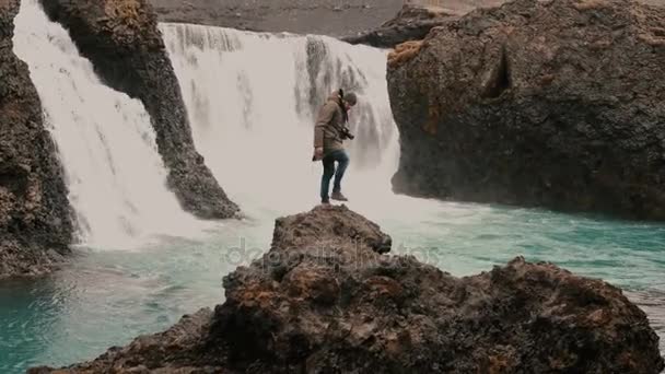 Young handsome man with camera walking alone in mountains valley near the powerful waterfall in Iceland. — Stock Video