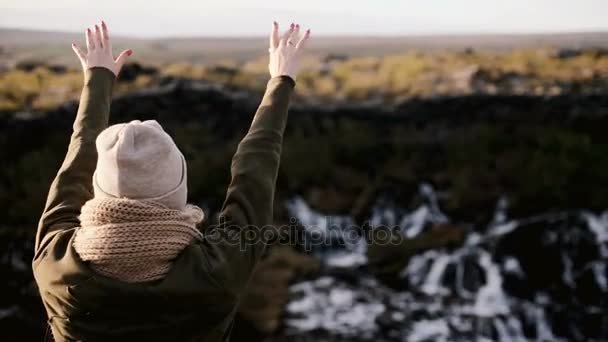 Mujer feliz de pie cerca de la cascada de Barnafoss en Islandia y saltando. Mujer joven se quita el sombrero y levanta la mano . — Vídeos de Stock
