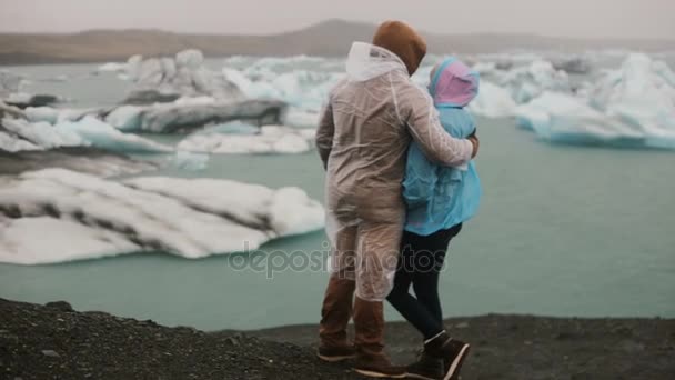 Achteraanzicht van jonge paar in regenjassen staande op de oever en kijken op gletsjers, ijsbergen in IJsland. — Stockvideo