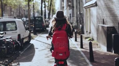 Arkadan Görünüm. Kadın bir güneşli şehir Bisiklete binmek. Ağır çekim. Bisiklet üzerinde kırmızı sırt çantası ile şık şapkalı kız. Keşfetmek.