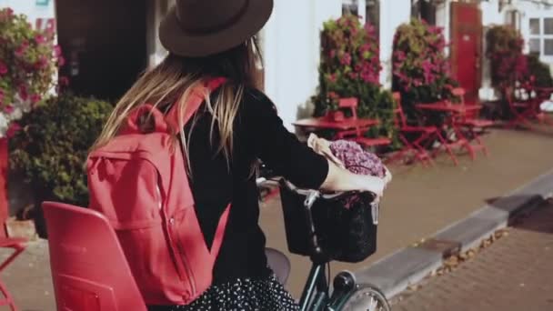 Chica sonriente monta una bicicleta con una mochila roja. Primer plano en cámara lenta. Mujer europea pasea por la calle con flores . — Vídeo de stock