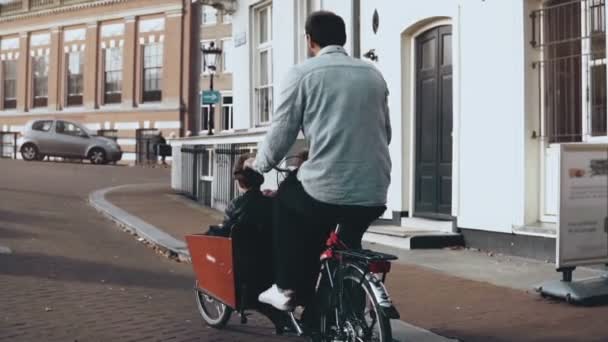 Hombre montando una bicicleta de carga con dos niños. Vista trasera en cámara lenta. Familia paseo en bicicleta feliz. Papá soltero. Madre soltera . — Vídeos de Stock