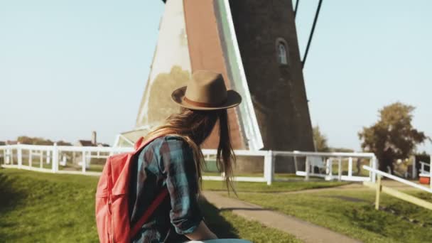 美丽的欧洲旅游女孩在老风磨房附近。美丽的年轻妇女在帽子与长发坐在村庄磨房附近。4k — 图库视频影像