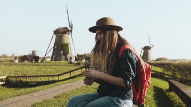 La joven local se sienta cerca de un viejo molino rústico. Vaquera en sombrero con pelo largo y mochila roja hace un ramo de flores. 4K — Vídeo de stock