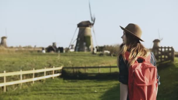 Lokale jong meisje loopt rond oude windmolen boerderij. Cowgirl in muts met lang haar en rode rugzak dwaalt zorgvuldig. 4k — Stockvideo