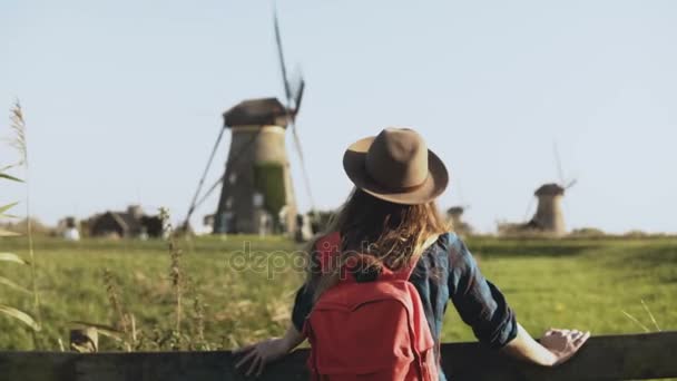Местная кавказка стоит рядом с ветряной мельницей. Деревенская женщина в шляпе с длинными волосами и красным рюкзаком оглядывается. 4K — стоковое видео