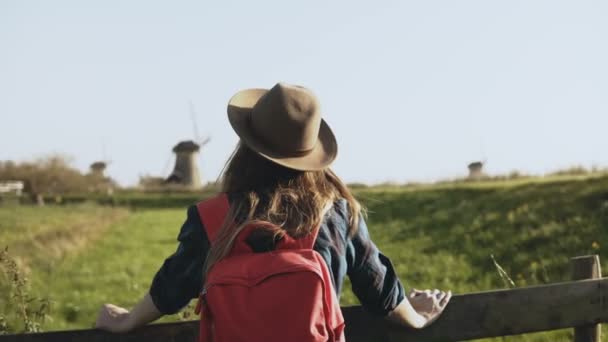 Портрет европейской девушки возле деревенской ветряной мельницы. Привлекательная женщина смотрит вокруг, наслаждаясь солнечным светом и счастливо улыбаясь. 4K — стоковое видео
