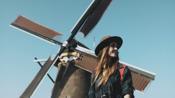 Portrait of traveller woman near old windmill. Incredible cinematic low angle. Girl in hat in front of farm mill. 4K — Stock Video