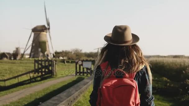 Chica local camina lentamente hasta una granja de molinos de viento. Mujer con sombrero, pelo largo y mochila roja volviendo a casa. 4K . — Vídeos de Stock