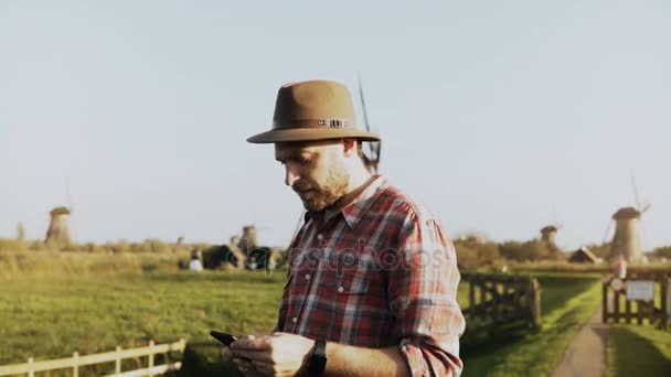4K Retrato de un granjero europeo exitoso. Hombre con camisa y sombrero, con smartphone y reloj inteligente camina a un molino . — Vídeo de stock