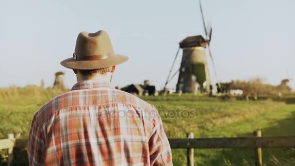 4 k úspěšné kavkazských větrný mlýn farmář dělník. Muž v košili dřevorubec a klobouk, rozhlédl se kolem sebe v sunny louka. — Stock video