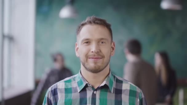 Retrato de un joven empresario exitoso en una oficina ocupada. Hombre guapo empleado mirando a la cámara y sonriendo. lento mo — Vídeos de Stock