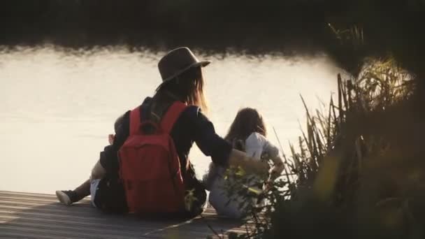 Γυναίκα, μικρό κορίτσι και αγόρι κάθονται στην προβλήτα στο ηλιοβασίλεμα. Εκπληκτικό κινηματογραφικό πλάνο. Ευτυχία. Έννοια της συντροφικότητας και της ενότητας. 4k. — Αρχείο Βίντεο