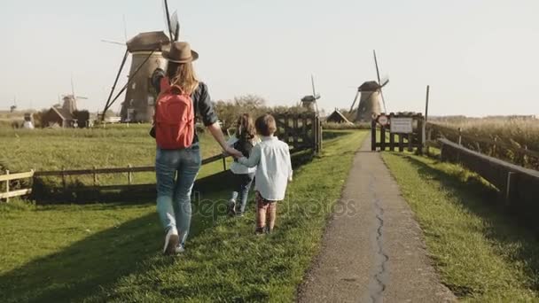 Yel değirmeni farm yakınındaki iki çocukla rahat anne. Kırmızı sırt çantası, küçük oğlan ve kız birlikte yürüyüş ile şapkalı kadın. 4k — Stok video