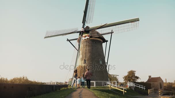 Familie van vier stand door enorme oude Nederlandse windmolen. Geweldig schot van mensen vóór een mijlpaal van het platform. Achteraanzicht. 4k. — Stockvideo