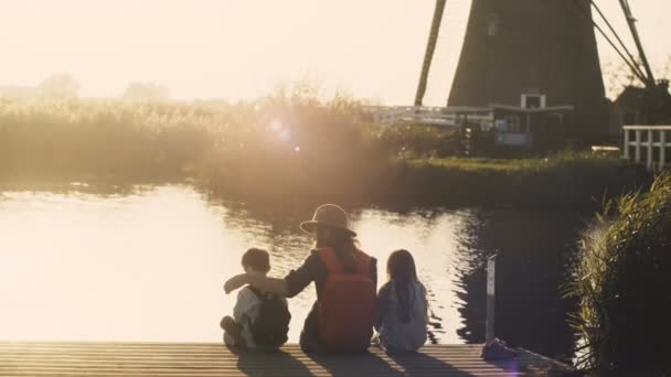 Woman with two kids sit on a lake pier on sunset. Amazing shot of a family together near water edge. 4K back view. — Stock Video